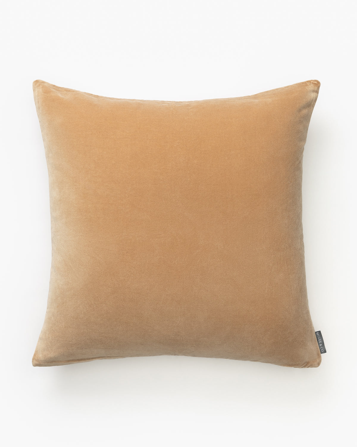 The Bellevue Velvet Pillow Cover, in a square, light brown stone-washed design with a soft texture, is shown on a white background. A small black fabric tag is attached to one side of the cover, by McGee & Co.