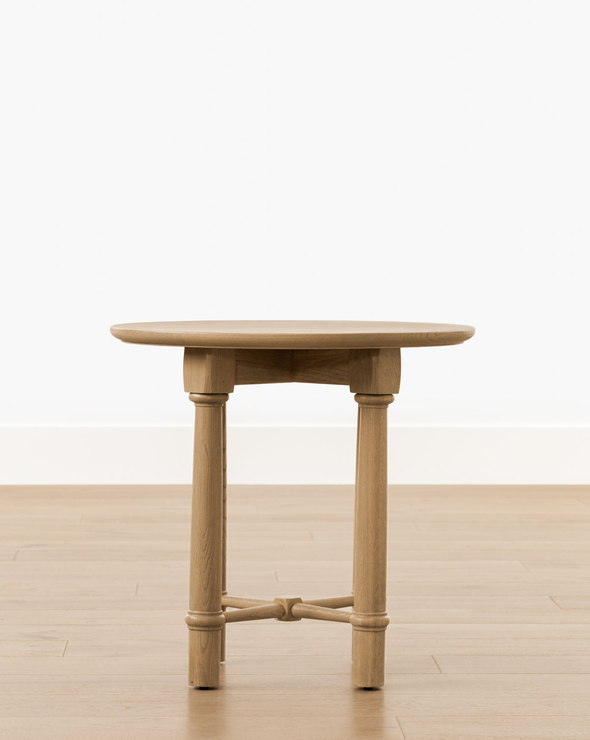 The Bishop Side Table features a round top, three turned wood legs, and a simple crossbar support, by McGee & Co. Shown here on a wooden floor with a plain white wall backdrop.