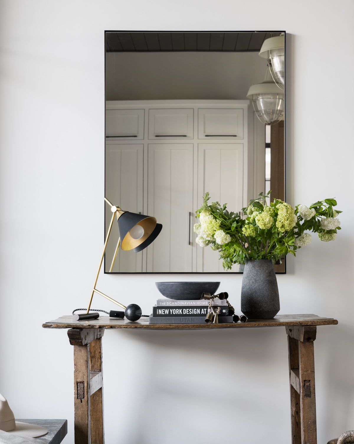 A rustic wooden table displays a modern black and gold desk lamp, stacked books, a grey vase with green and white flowers, a bowl, and against a white wall hangs the Knowles Mirror with its sleek stainless steel frame, by McGee & Co.