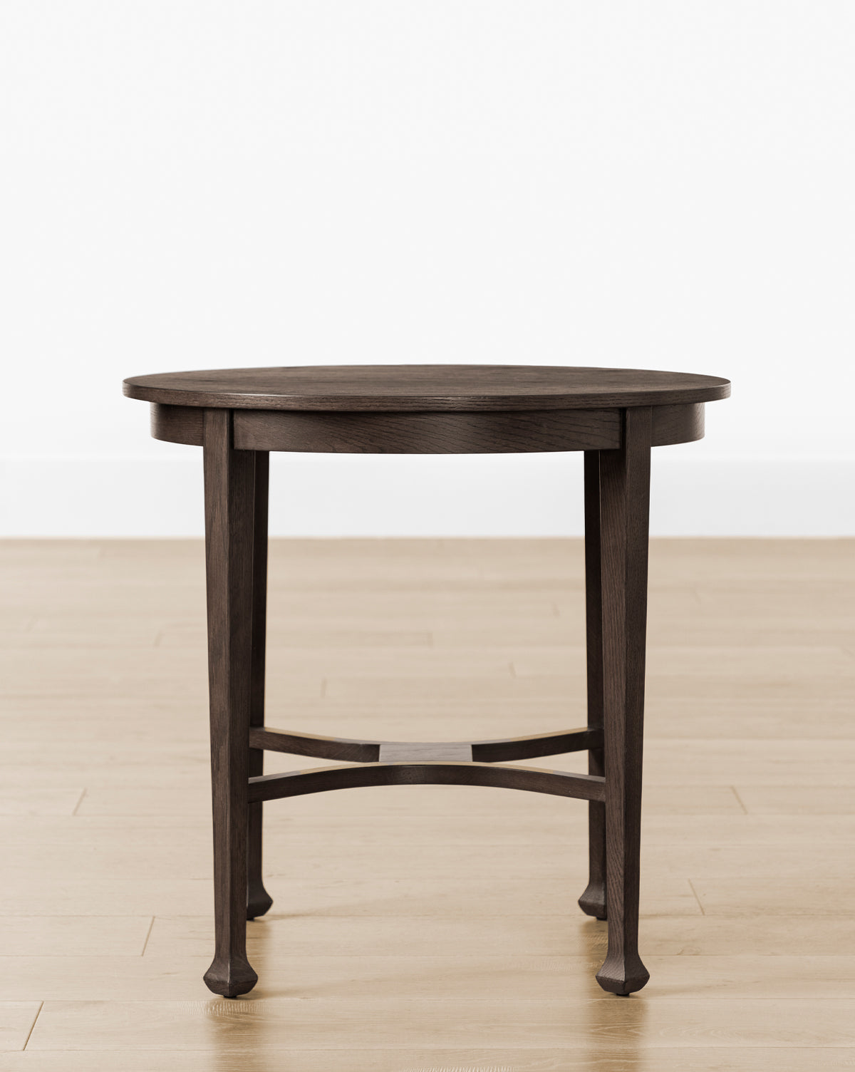 The Bradshaw Side Table, featuring a round dark wood top with a satin walnut oak finish and an antique-style crossbar base, stands on a light wood floor against a plain white wall, by McGee & Co.