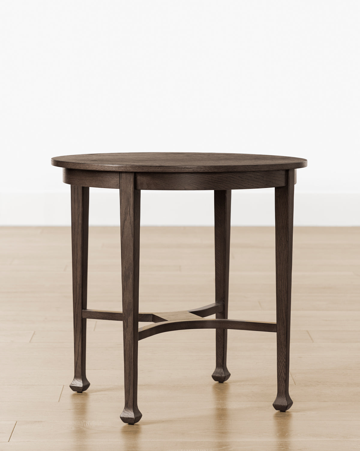 The Bradshaw Side Table showcases a round, dark satin walnut oak finish with four legs and a crossbar support, displayed on a light wood floor against a plain white wall, by McGee & Co.