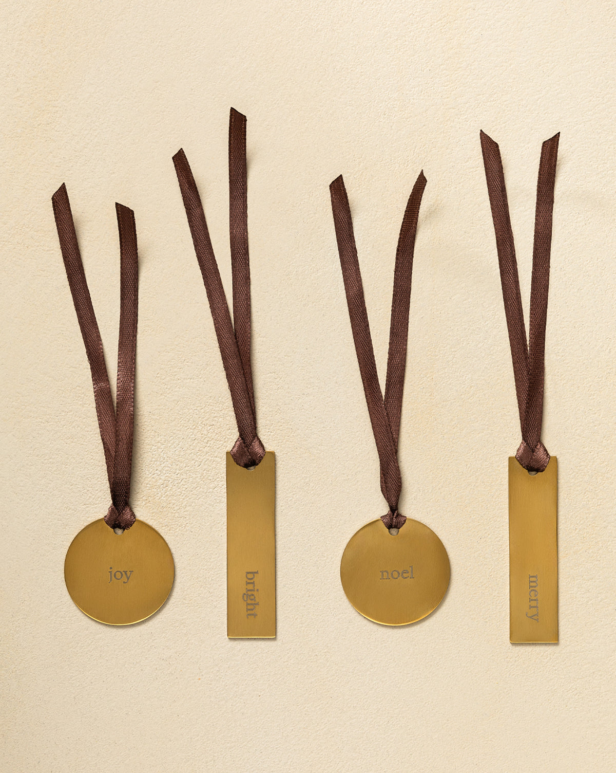 Four Brass Holiday Gift Tags with brown ribbon hang against a beige background—two round tags read “joy” and “noel,” while two rectangular tags say “bright” and “merry,” by McGee & Co.