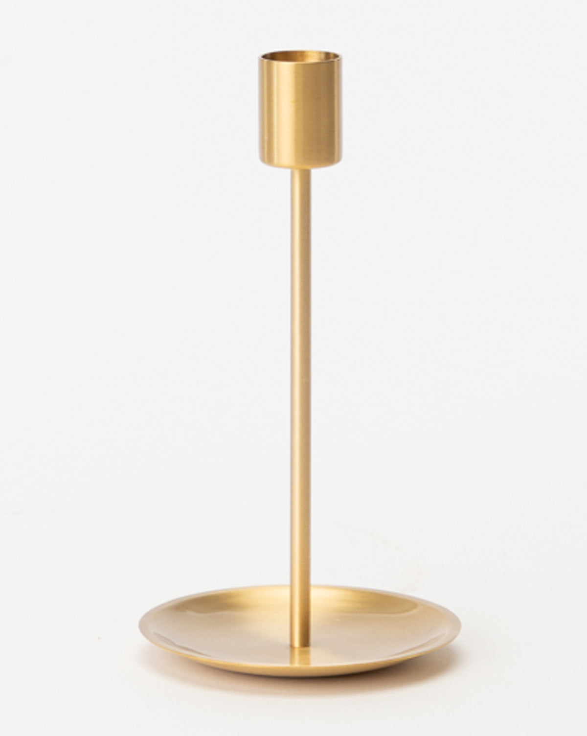 The Brass Taper Holder showcases a minimalist gold-tone metal design with a round base, slender stem, and cylindrical top candle holder, all set against a plain white background.