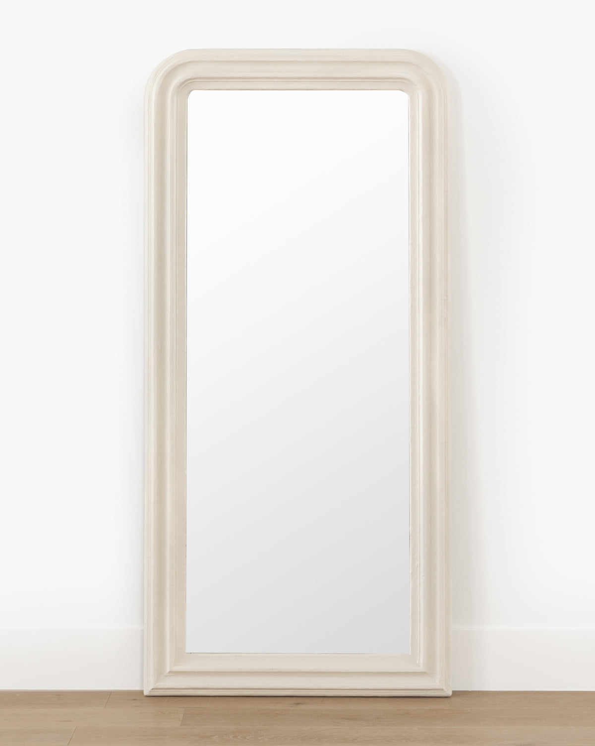 The Breslin Floor Mirror, featuring a tall rectangular shape with a rounded top and thick light-toned frame, stands against a white wall on a light wood floor, displaying its elegant plaster finish, by McGee & Co.