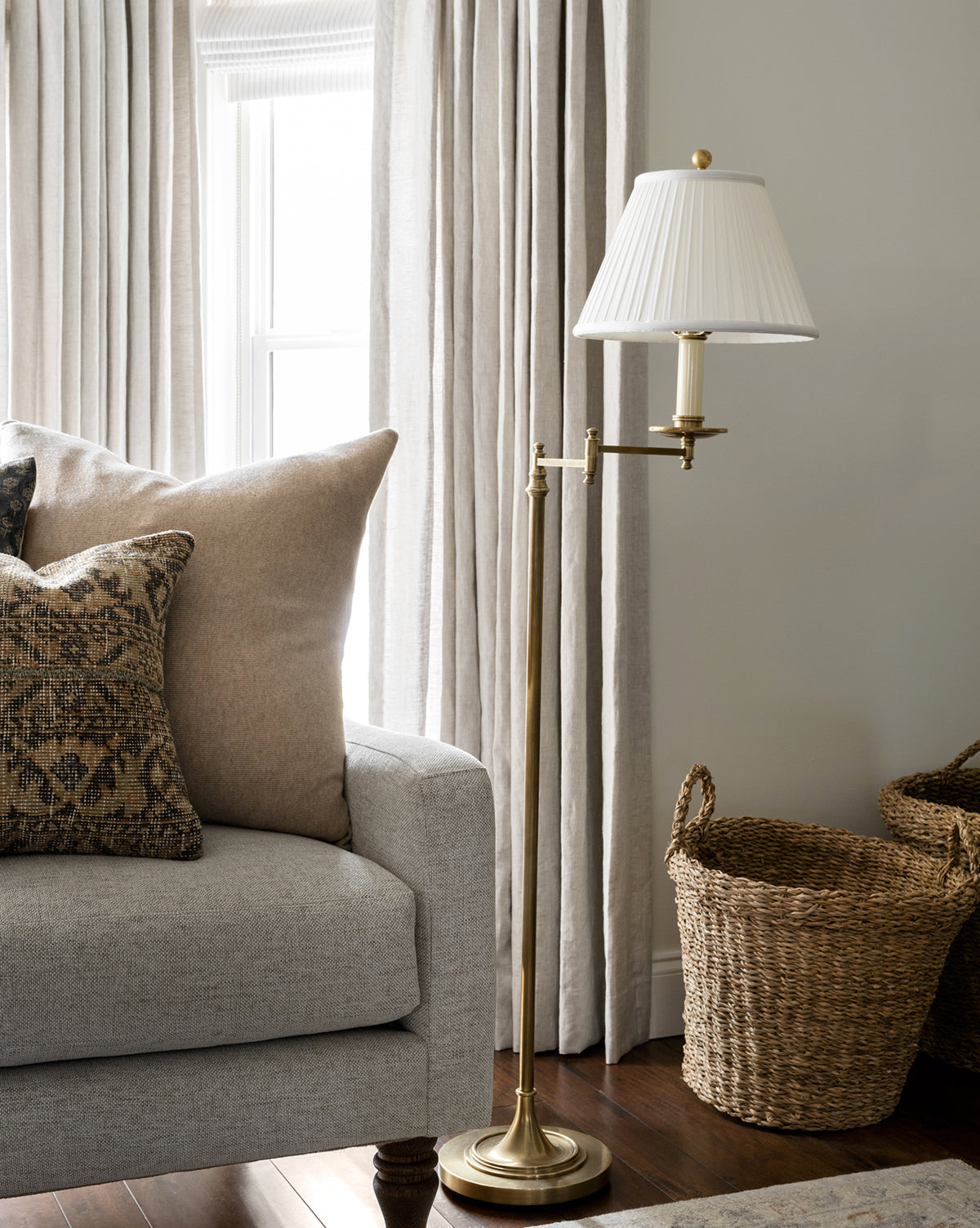 The Dorchester Swing Arm Floor Lamp with a pleated silk shade stands beside a light gray sofa topped with patterned and solid throw pillows. Two woven baskets sit on the wooden floor near beige curtains by the window, by McGee & Co.