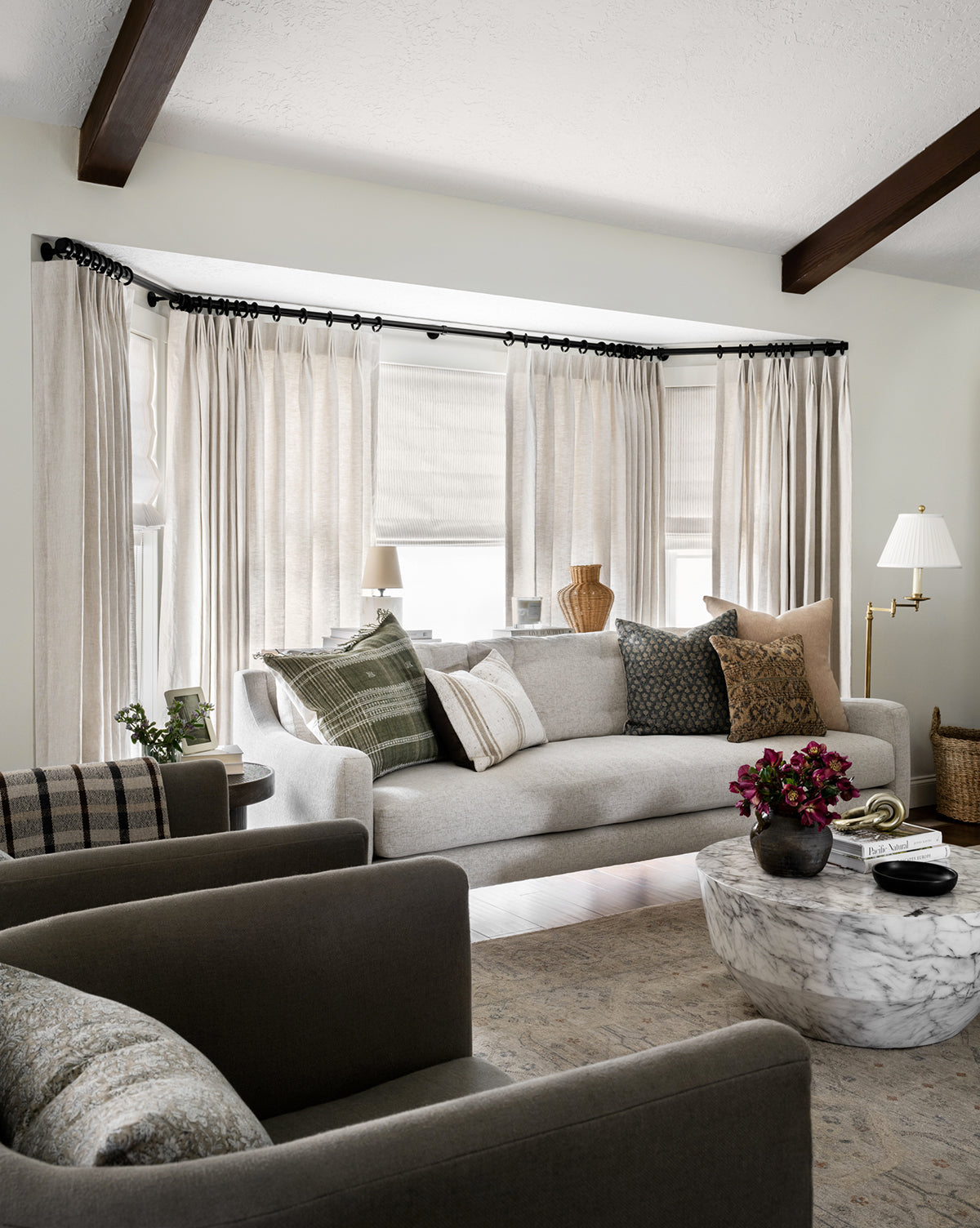 A contemporary living room with a Barden Sofa 84 featuring tufted cushions and reeded wood legs, patterned pillows, two dark armchairs, a round marble coffee table with flowers, sheer curtains, vases, a floor lamp, and exposed ceiling beams, by McGee & Co.
