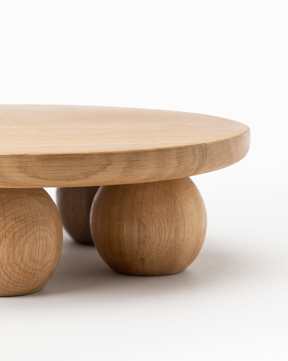 The Bronx Oak Pedestal is a minimalist, modern cake stand with four spherical legs and a smooth, light oak finish. Its round wooden tray design stands out against any plain background, making it perfect for contemporary presentations by McGee & Co.