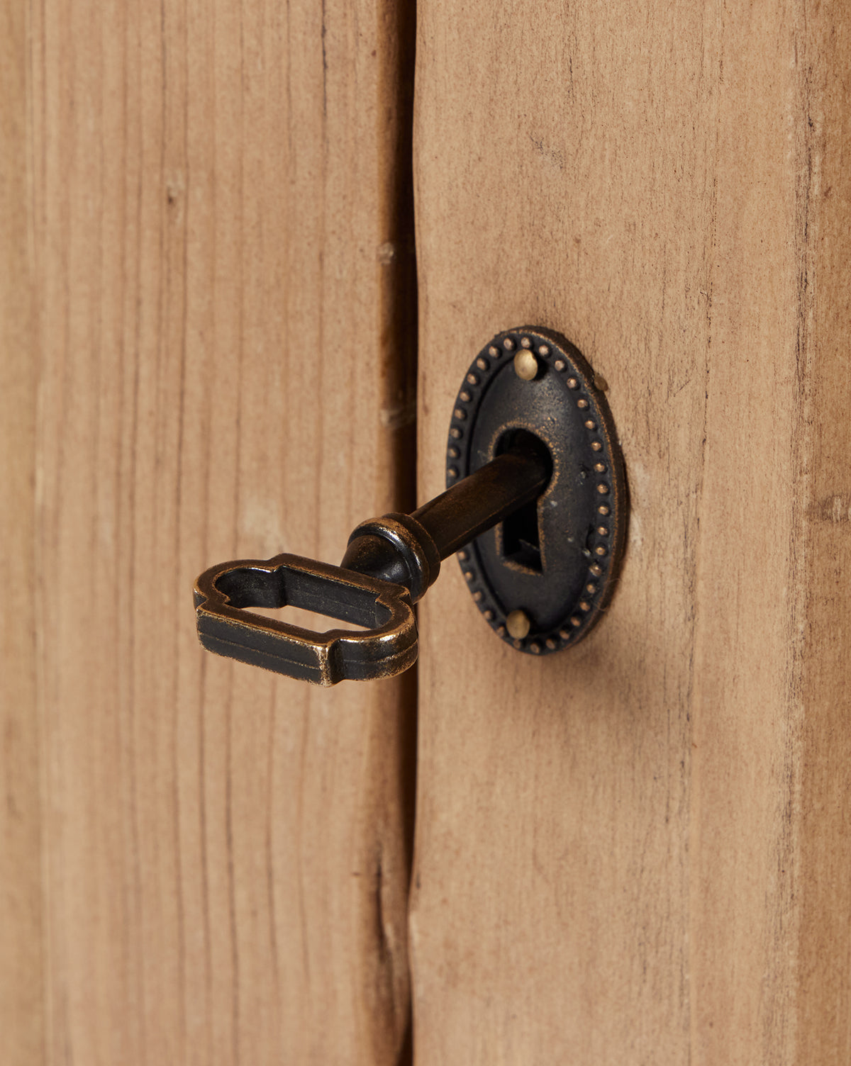 A vintage brass key enters the ornate keyhole of the Bryn Cabinet’s door, crafted from solid pine with a natural finish, by McGee & Co.