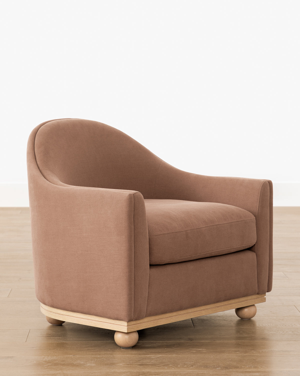 The Bunnell Lounge Chair (Ready to Ship) features a curved back, armrests, and light brown upholstery. Its modern traditional design sits on a wooden base with round feet, displayed on a light wood floor against a plain white wall by McGee & Co.
