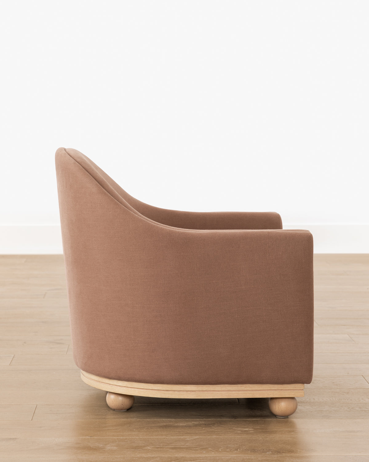 The Bunnell Lounge Chair (Ready to Ship) features modern brown upholstery, a rounded back and arms, and a light wood base with ball-shaped feet. Its shown on a wooden floor against a white wall by McGee & Co.
