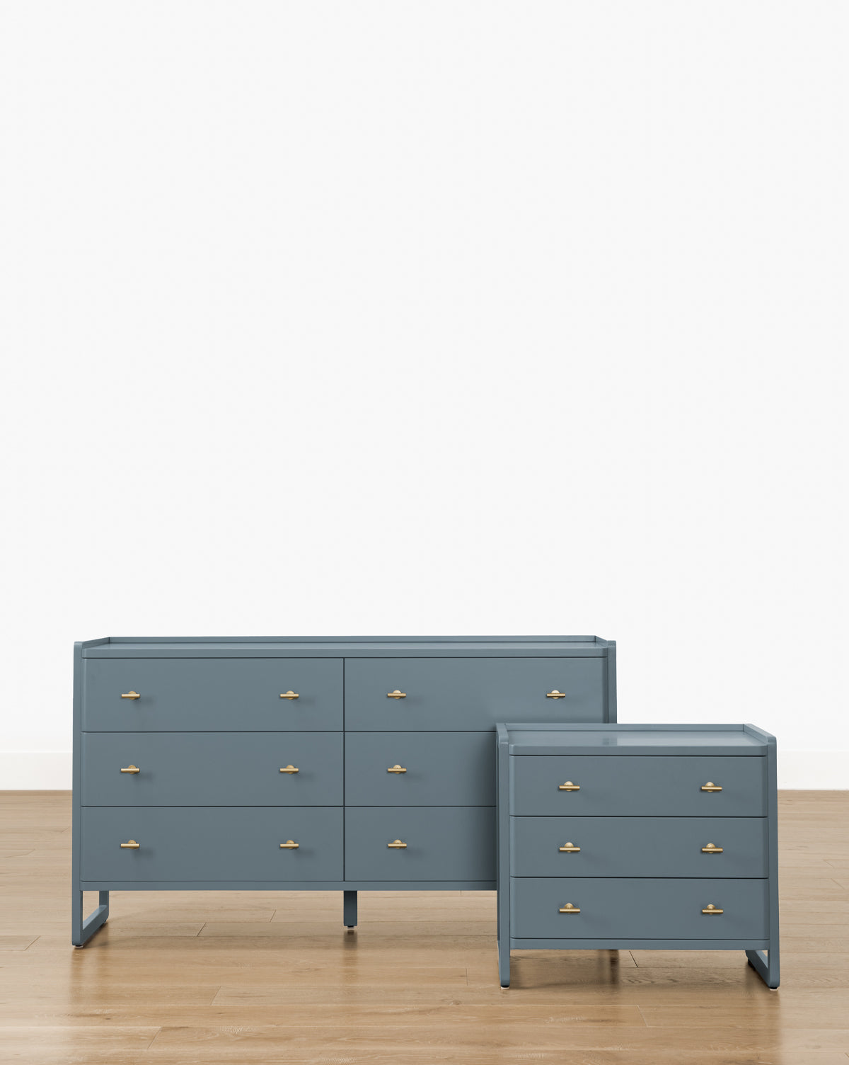 A classic-style Butler Nightstand in slate blue with gold handles stands beside a blue six-drawer dresser on a light wood floor, set against a plain white background by McGee & Co.