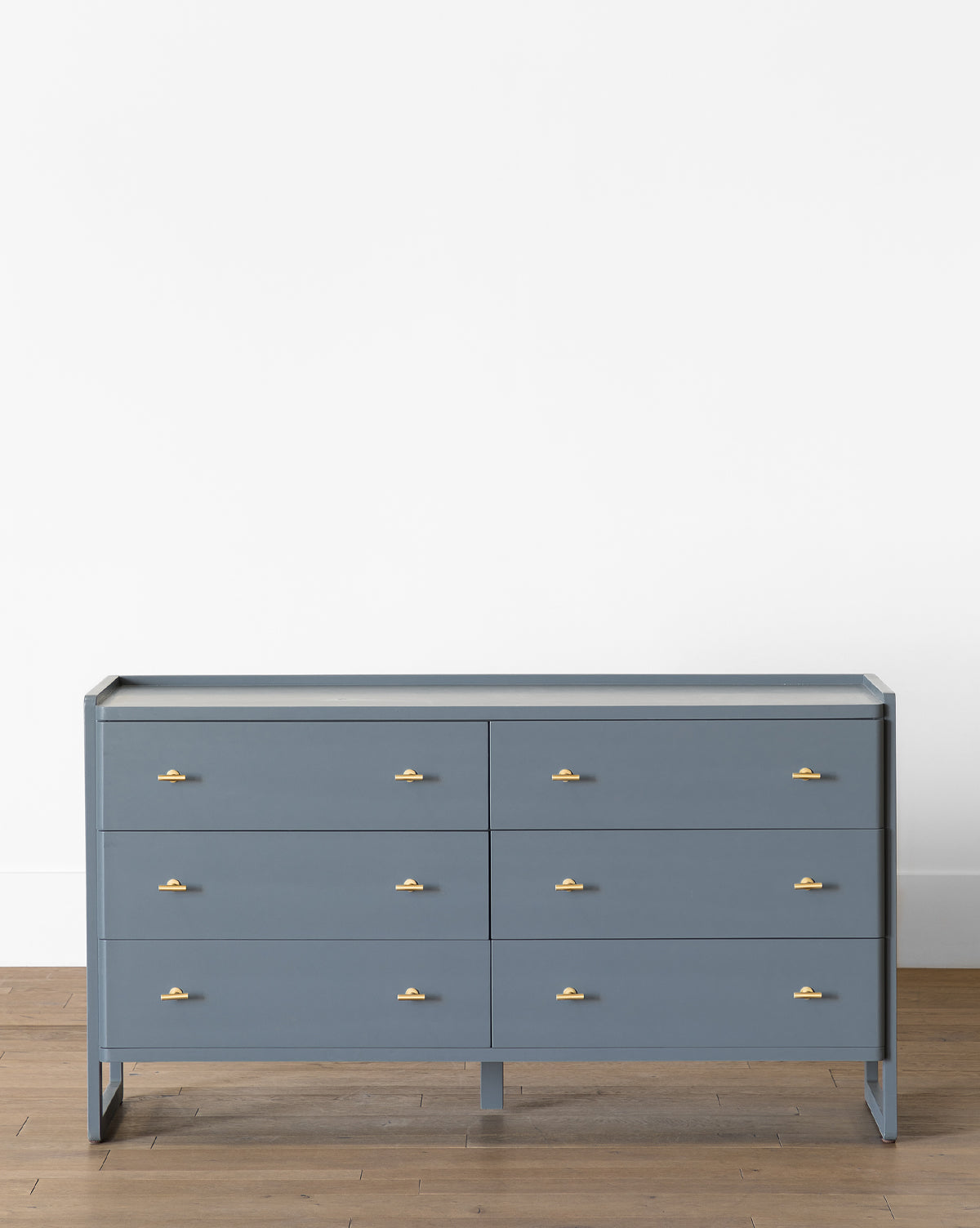 The Butler Dresser, in slate blue with six gold-handled drawers, stands on a wooden floor against a white wall, bringing elegance to any bedroom, by McGee & Co.