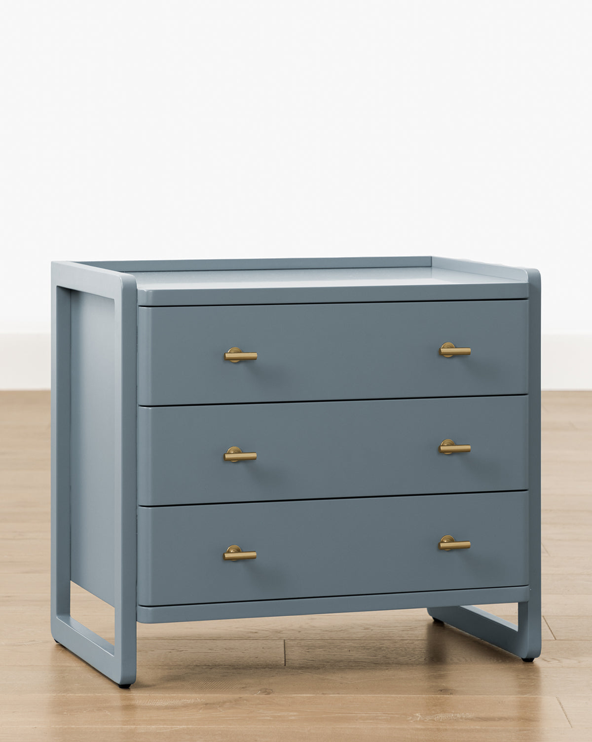 The Butler Nightstand, a modern slate blue piece with three drawers and sleek gold handles on angled legs, is displayed on a light wood floor against a plain white background by McGee & Co.