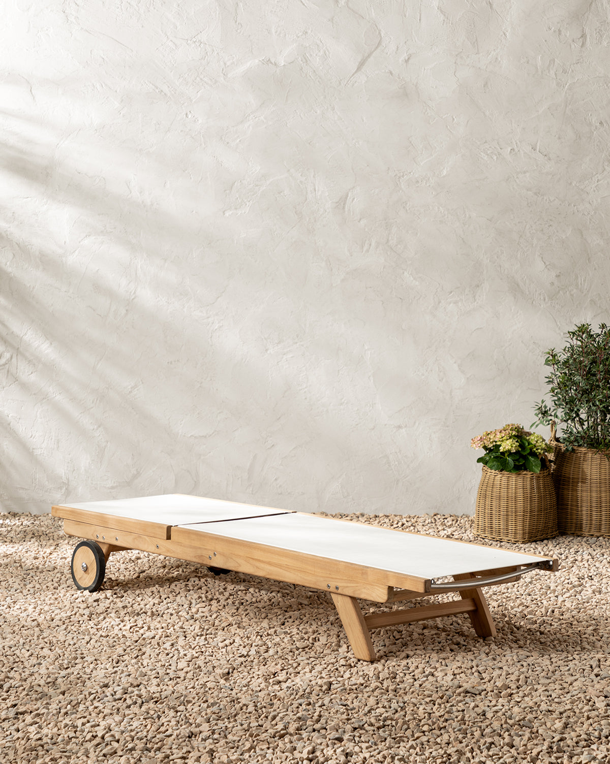 The Cadie Outdoor Chaise features white weather-resistant cushions and sits on gravel before a textured beige wall. Two woven baskets with green plants appear in the background on the right, by McGee & Co.
