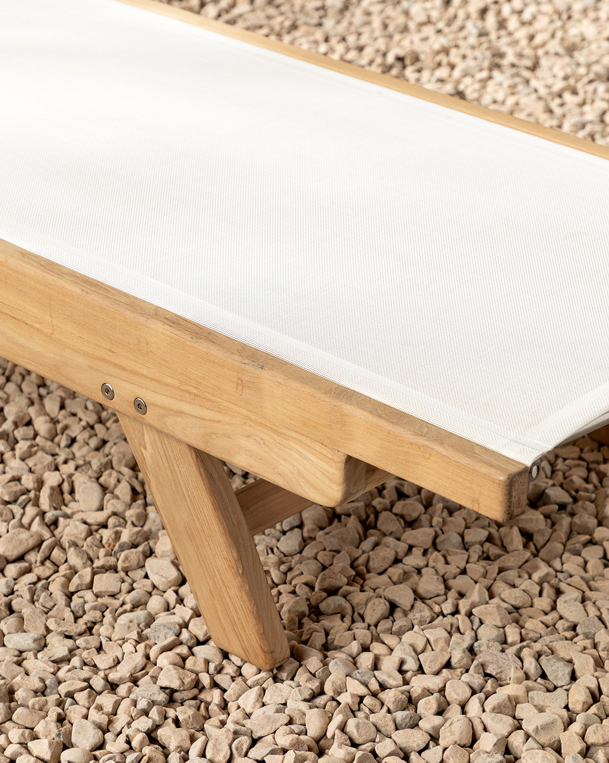 A close-up of the Cadie Outdoor Chaise, featuring a teak frame and white weather-resistant fabric seat, set on a bed of small, light-colored pebbles by McGee & Co.