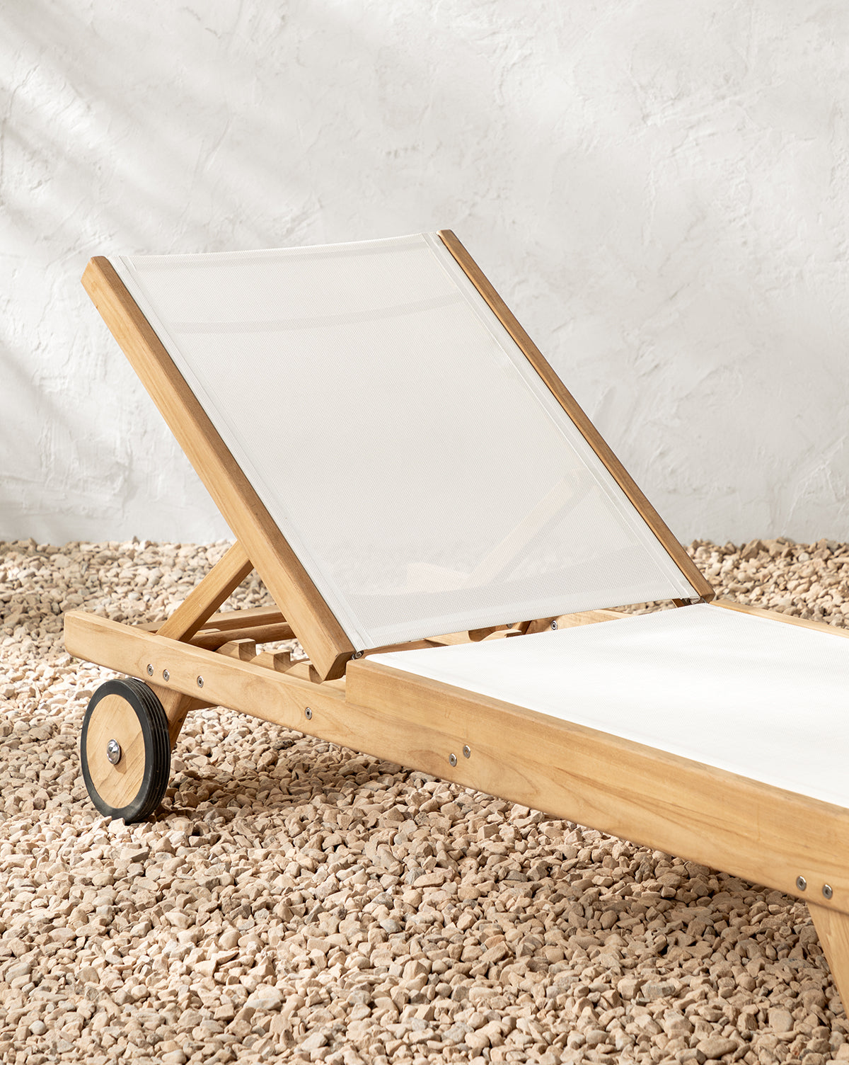 The Cadie Outdoor Chaise, featuring weather-resistant fabric on wood, rests on light gravel beside a textured white wall by McGee & Co.