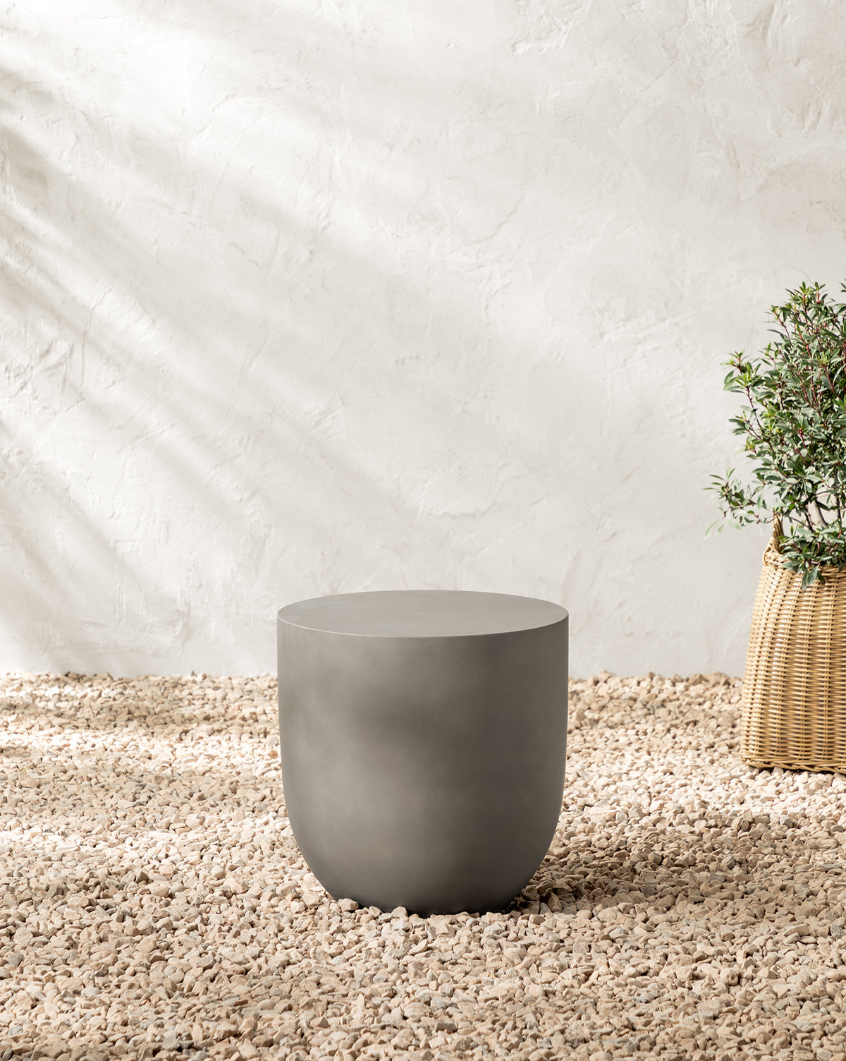 The Caine Outdoor Side Table stands on a pebble-covered floor by a plant in a woven basket, while sunlight casts soft shadows across the textured white wall, by McGee & Co.