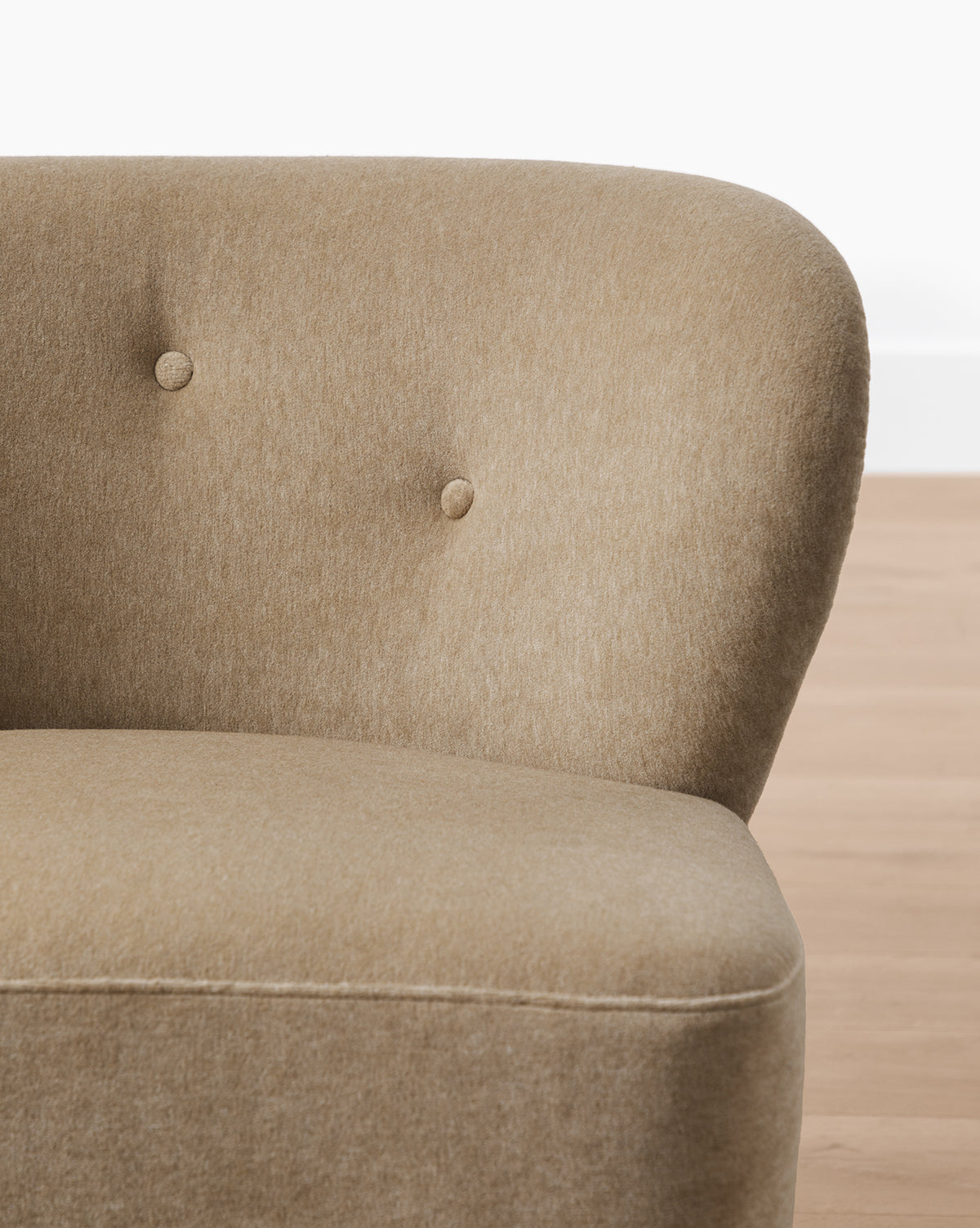 Close-up of the Carmella Slipper Chair (Ready to Ship) in beige with camel upholstery, a curved backrest, two fabric-covered buttons, and soft fabric. Set against light wooden flooring and a white wall, by McGee & Co.