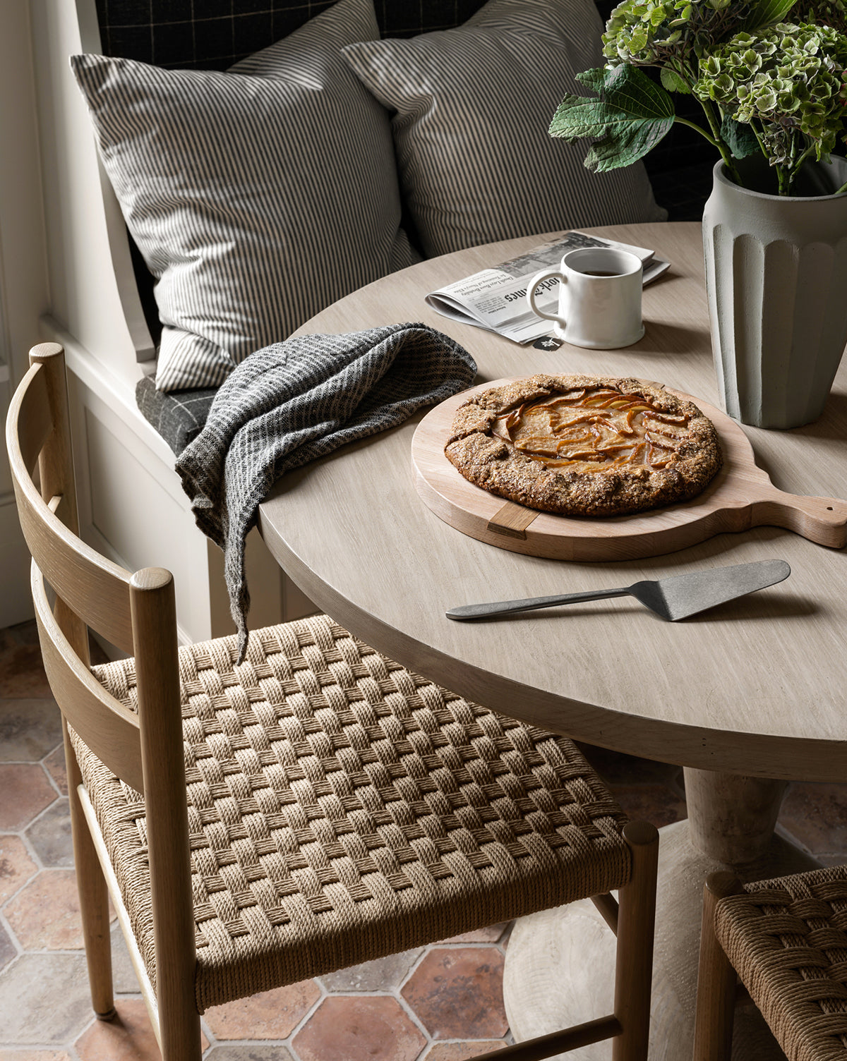 A cozy breakfast nook features Danish design with Eloise Woven Chairs and a round wooden table topped with a rustic fruit galette, pie server, mug, newspaper, and green hydrangeas. Pillows and a blanket add comfort, by McGee & Co.