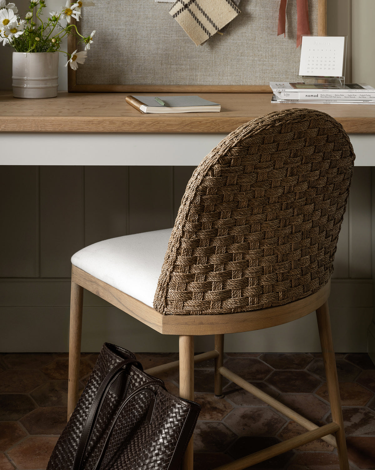A Molly Dining Chair with a white cushion is placed by a wooden desk with notebooks, a calendar, plant, and decor. A woven brown bag sits on the floor next to this modern Molly Dining Chair by McGee & Co.