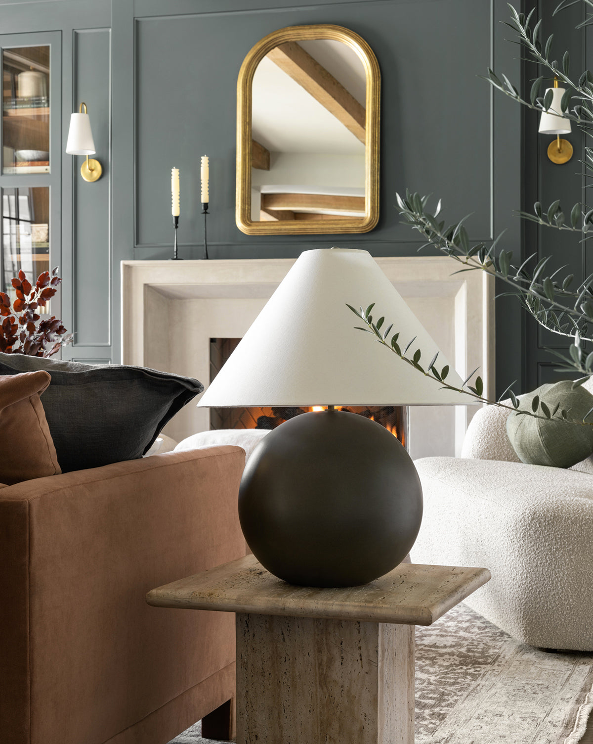A modern living room features a brown sofa, cream armchair, and the Tess Large Ceramic Table Lamp on a stone side table. A fireplace with a gold-framed mirror, plus candles and greenery, complete the stylish decor by McGee & Co.