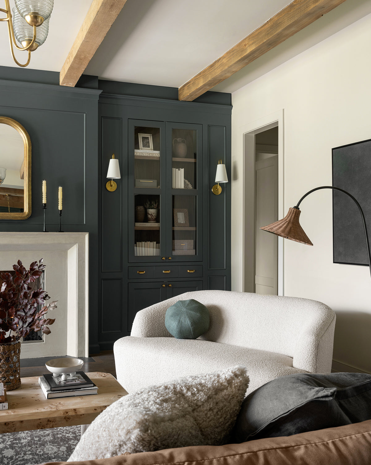 A modern living room features the Makerspalm Byron Settee 68 with a curved back, a wooden coffee table, and built-in dark cabinets with glass doors. Decorative objects, books, and soft lighting add to the cozy, stylish space. McGee & Co.