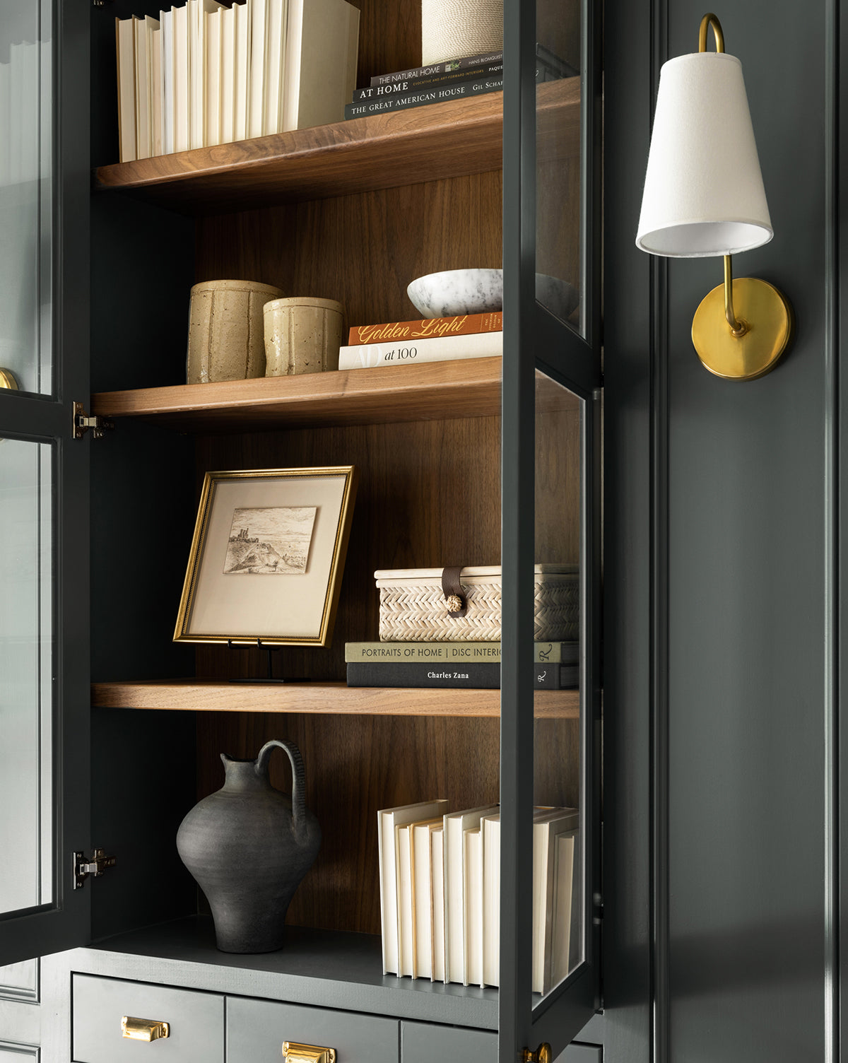 A glass-fronted cabinet with wooden shelves displays books, ceramic vases, a framed sketch, decorative boxes, and the Charcoal Terracotta Jug. Beside the cabinet is a wall sconce with brass and white accents.