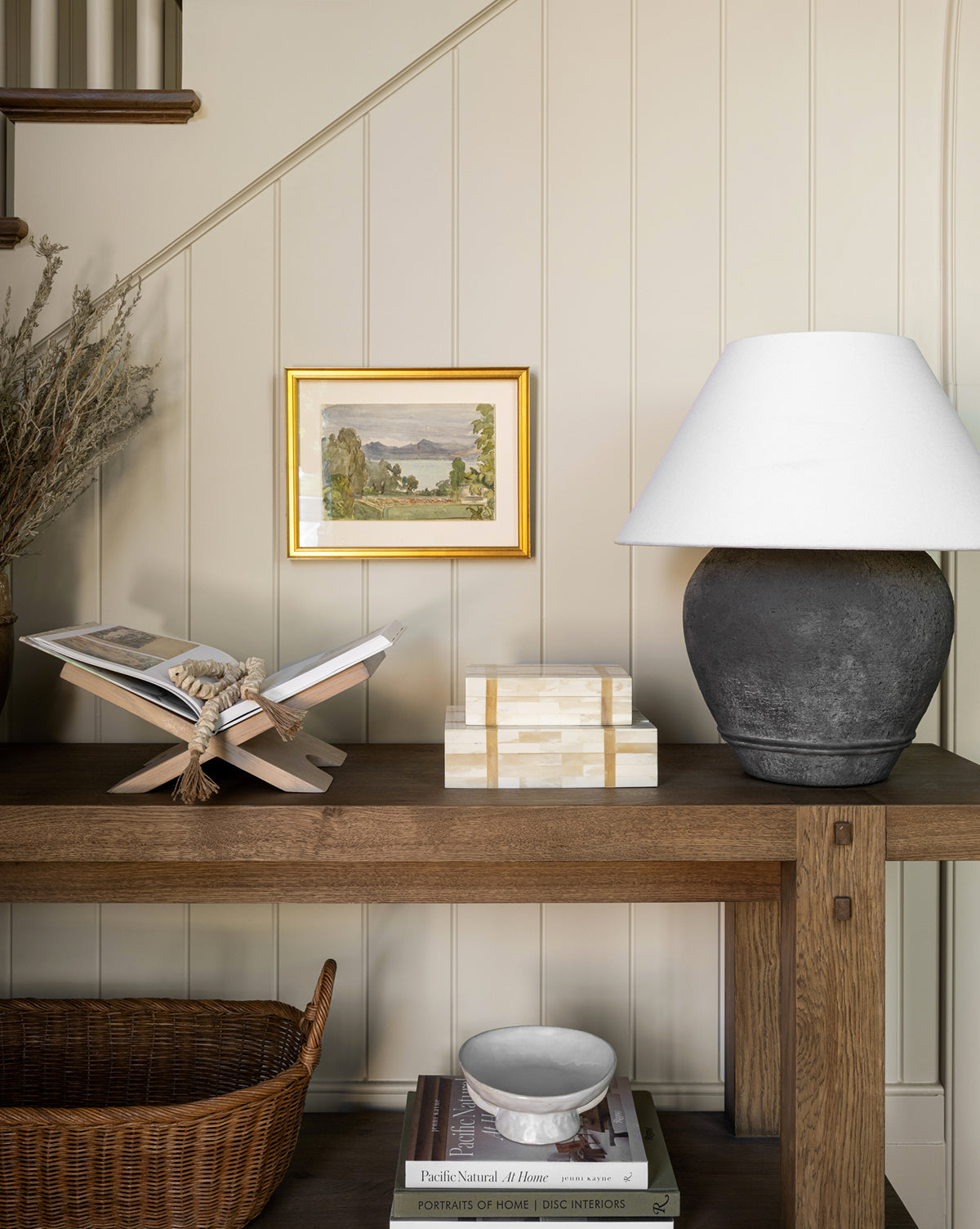 A wooden console table holds a dark lamp with a white shade, a decorative box, an open book on the Wooden Book Display, and a framed landscape painting on a beige paneled wall. A wicker basket sits below the table.