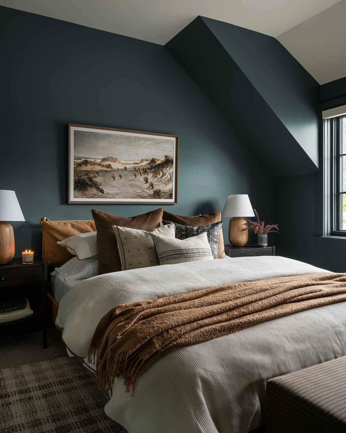 A cozy bedroom with dark teal walls, a large bed with tan and cream bedding, brown throw blanket, multiple pillows, two bedside tables with lamps, a candle, and the Morning Sand Dune giclee print in a wood frame above the bed.