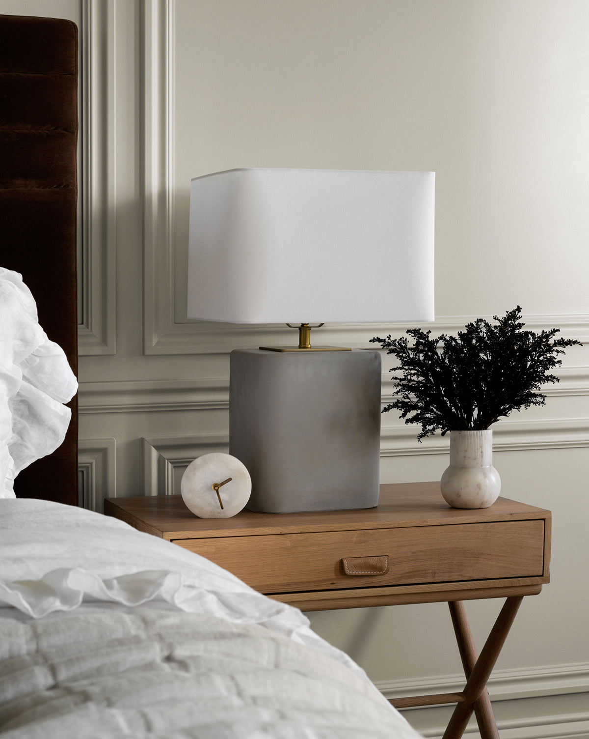 A wooden bedside table with an Alabaster Table Clock, a gray square lamp, and a white vase of dark foliage stands beside a bed with white linens. Paneled walls and a brown headboard contribute to the sophisticated design.