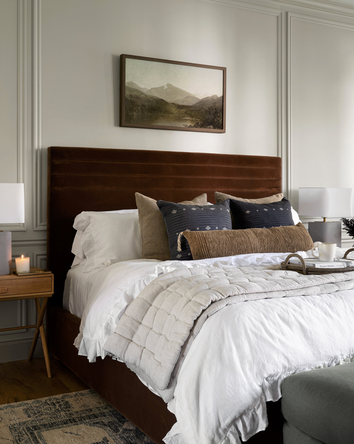 A cozy bedroom features a brown upholstered bed with white bedding, decorative pillows, and a wooden nightstand topped with a candle and lamp. The Darsey Lake Peaks giclee print hangs above the bed, and a breakfast tray sits at the foot.