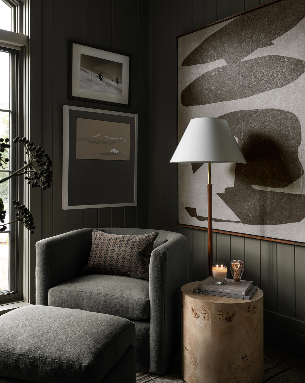A cozy, modern reading nook featuring a gray armchair, leather ottoman, side table with candle and glass, and the Elijah Floor Lamp with a brass pole—set against dark-paneled walls with abstract art by a sunlit window by McGee & Co.
