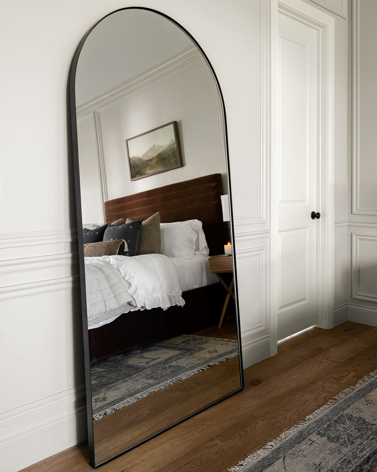 The Gala Floor Mirror with a black frame leans against a white wall, reflecting a cozy bedroom with a bed, pillows, wall art, and nightstand. Light wood floors and a patterned rug add warmth to the inviting space, by McGee & Co.