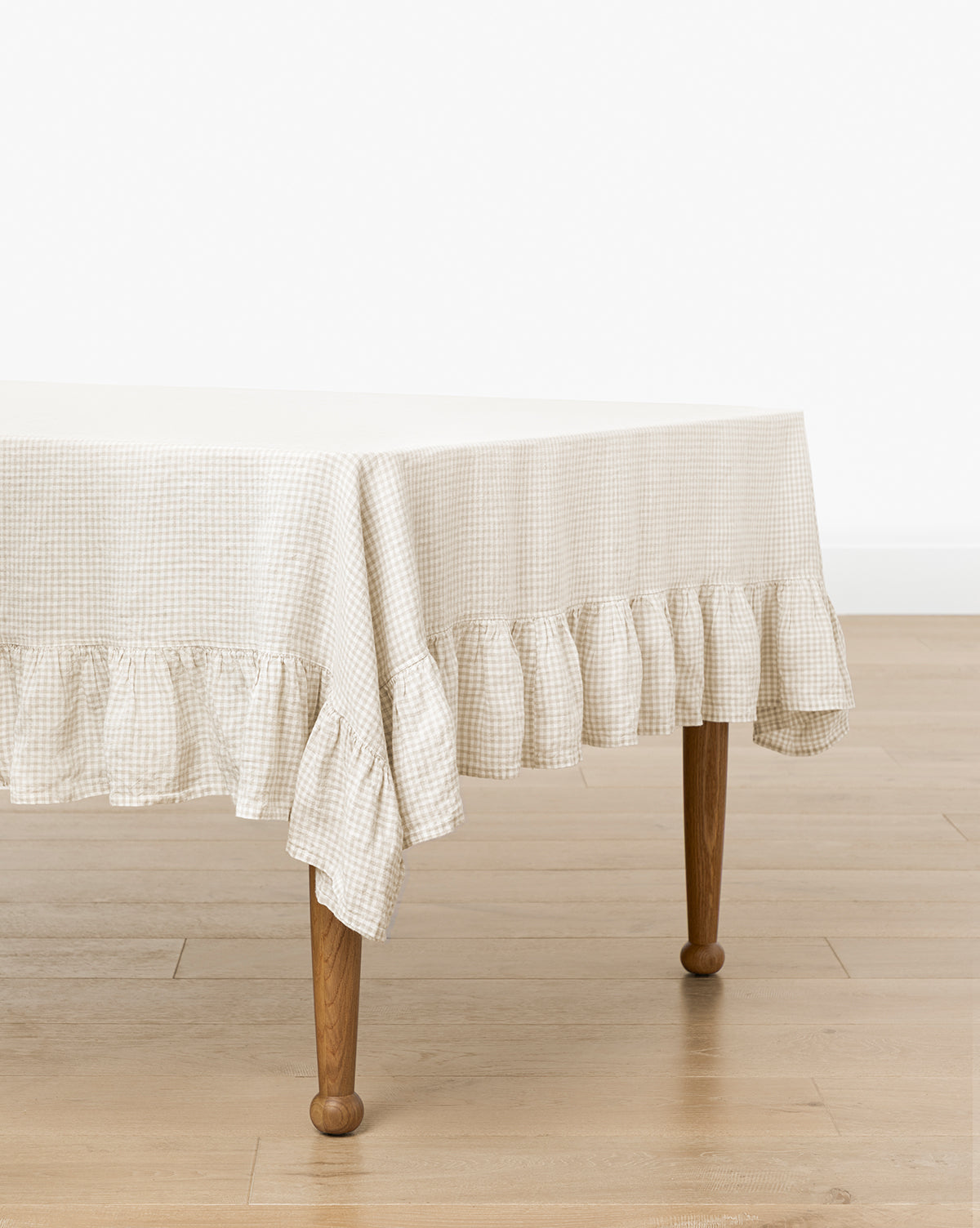 A rectangular table is covered with the Catania Linen Ruffle Tablecloth in beige gingham, featuring a ruffled edge, set on a light wooden floor against a white wall, by McGee & Co.