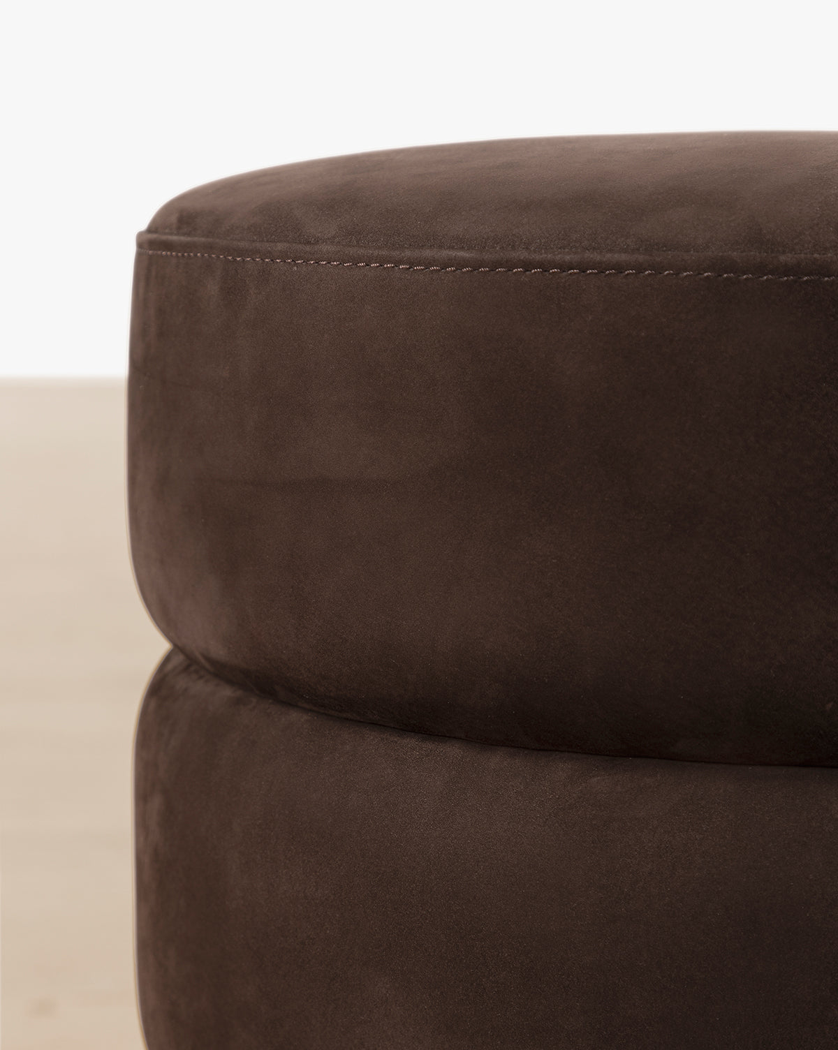 Close-up of the Channel Ottoman, a round, dark brown pouf with quilted suede-like upholstery, showcasing its soft stitched surface and subtle midcentury style against a neutral background, by McGee & Co.