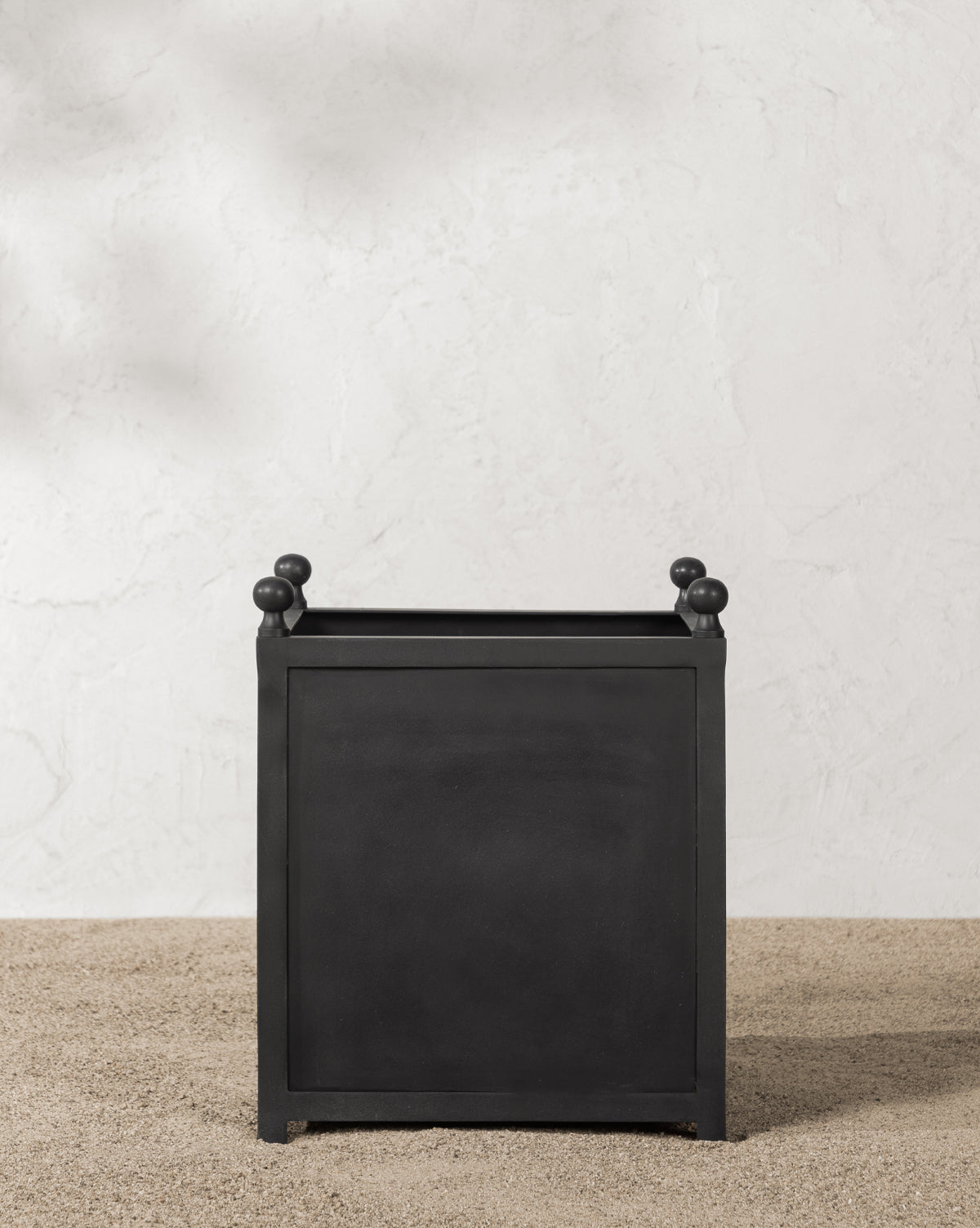 The Clariance Planter, featuring a square shape with small rounded corner knobs, rests on light pebbles, making it ideal for outdoor decor against a textured white wall.