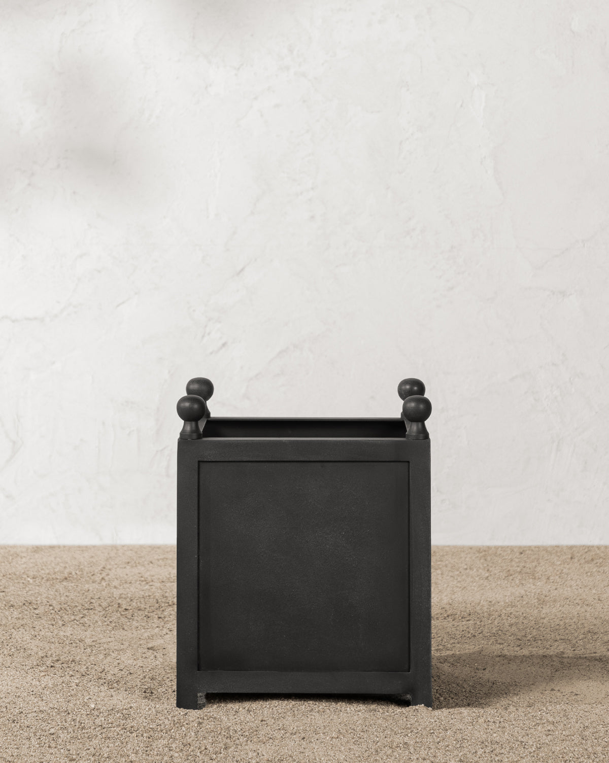 A black Clariance Planter with small rounded feet and decorative knobs rests on light pebbles against a textured white wall, bathed in sunlight—perfect for outdoor decor.