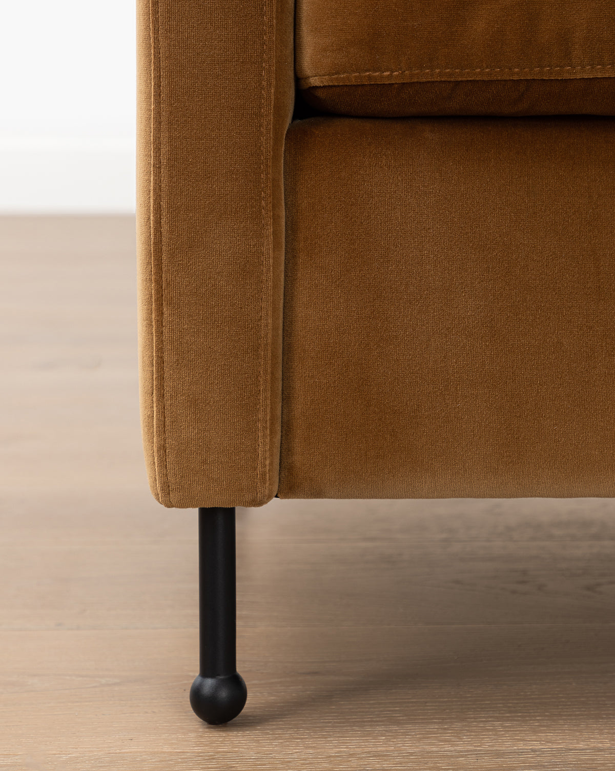 Close-up of the bottom corner of the Clegg Lounge Chair (Ready to Ship), featuring brown cotton velvet upholstery and iron legs, set on a light wooden floor against a white wall, by McGee & Co.