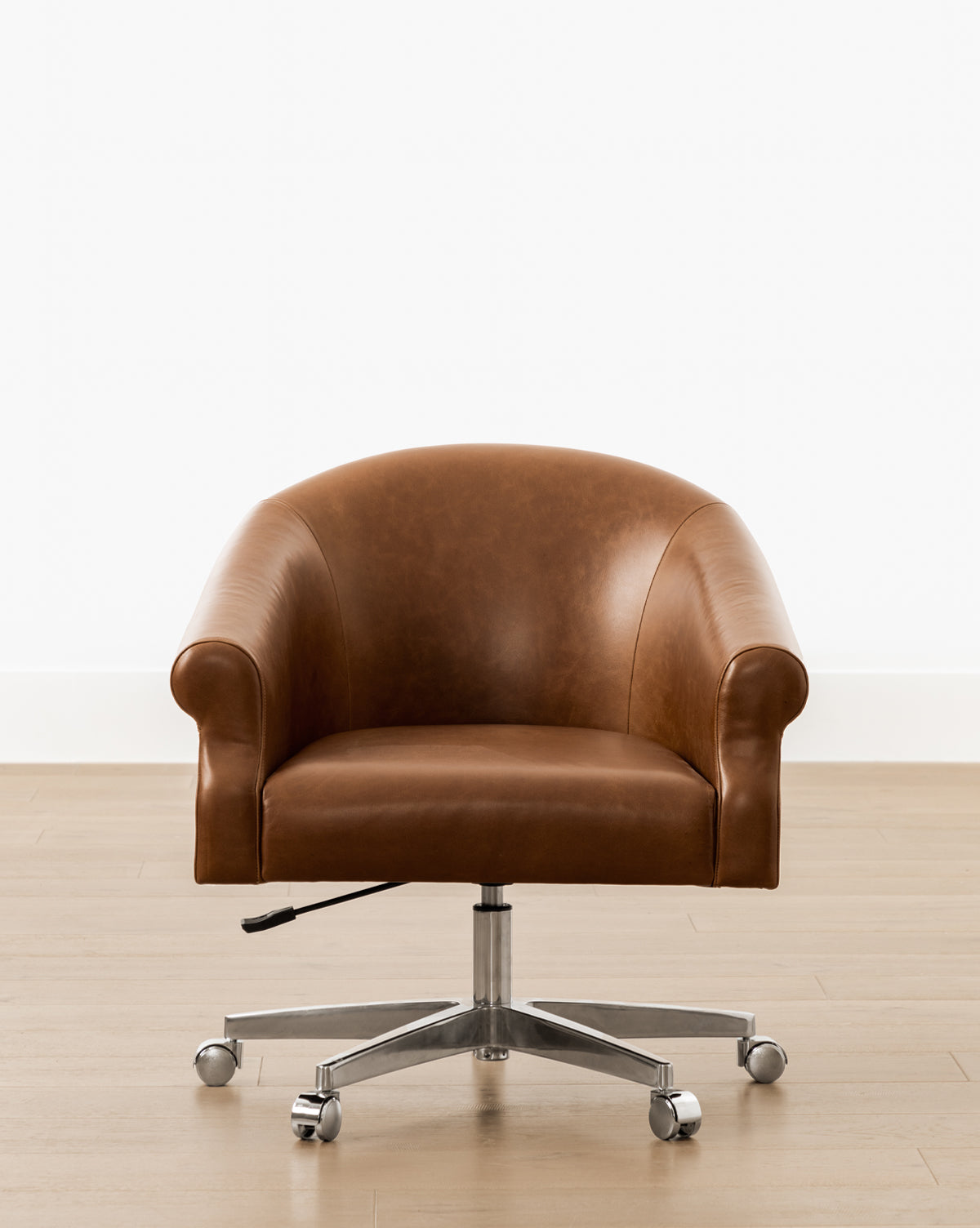 The Clemence Leather Desk Chair, featuring brown saddle leather, rounded armrests, and a metal base on casters, adds style to your home office. Shown here on a light wooden floor against a plain white wall by McGee & Co.