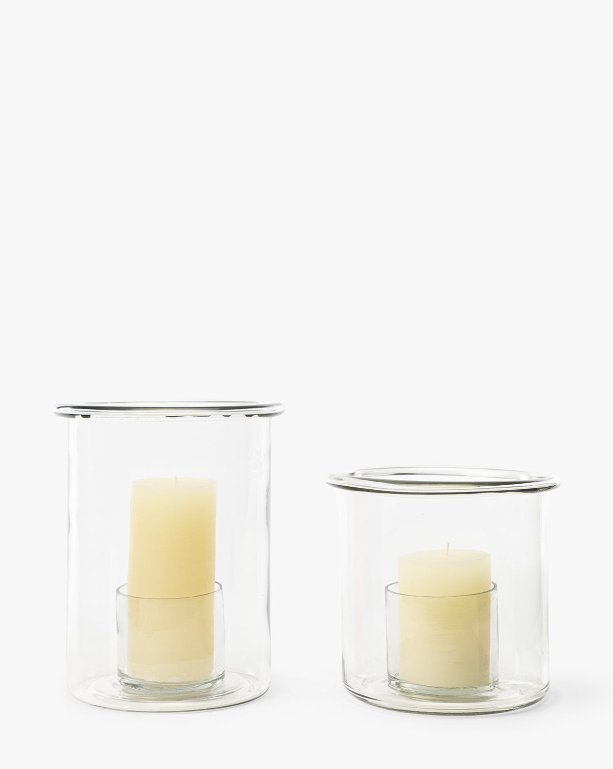 Two cream-colored pillar candles in Cyrus Hurricane clear glass holders, one tall and one short, cast soft ambient light against a plain white background by McGee & Co.