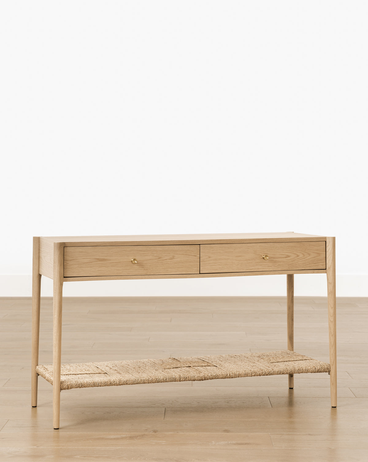 The Dana 2-Drawer Console features oak wood, two drawers with gold knobs, and a woven lower shelf, standing on a wooden floor against a plain white background—ideal for enhancing your entryway décor by McGee & Co.