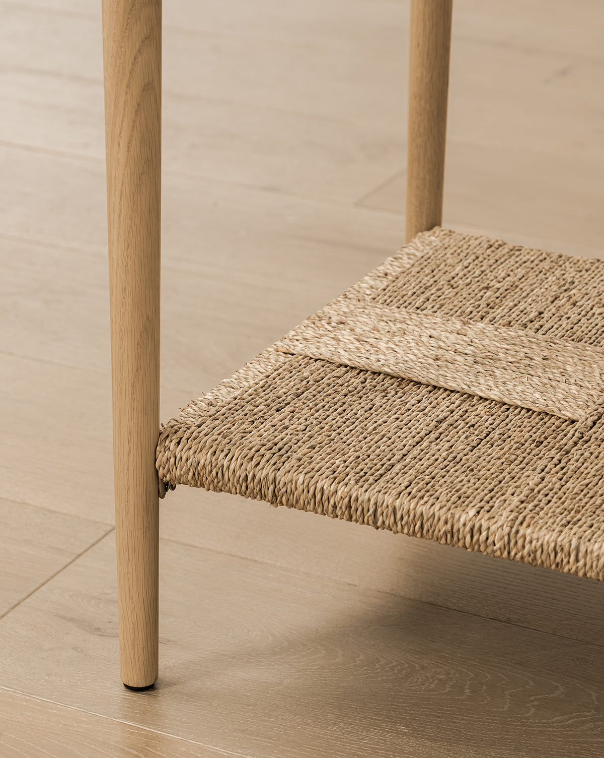 Close-up of the Dana 2-Drawer Console’s oak leg on a light wood floor, featuring a woven rattan shelf between the legs—a stylish choice for entryway decor, by McGee & Co.