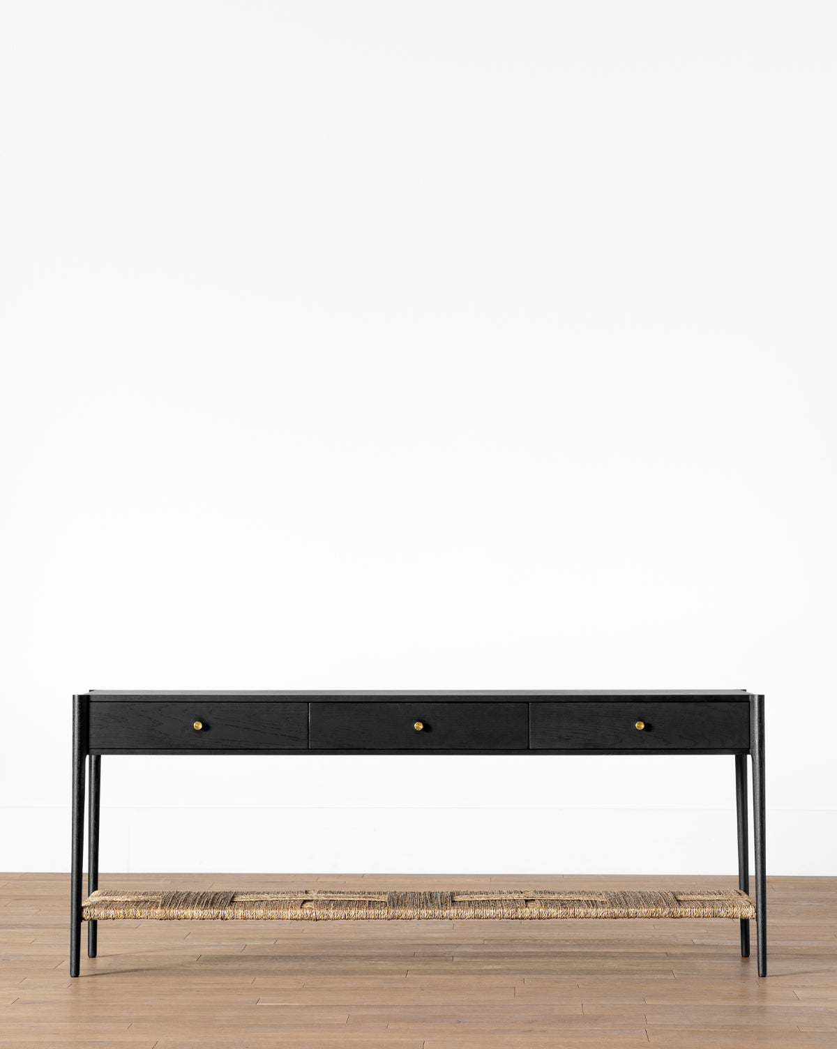The Dana Console is a minimalist black table featuring three drawers with gold knobs, slim legs, and a woven seagrass shelf—perfect entryway decor against a white wall on a wooden floor, by McGee & Co.