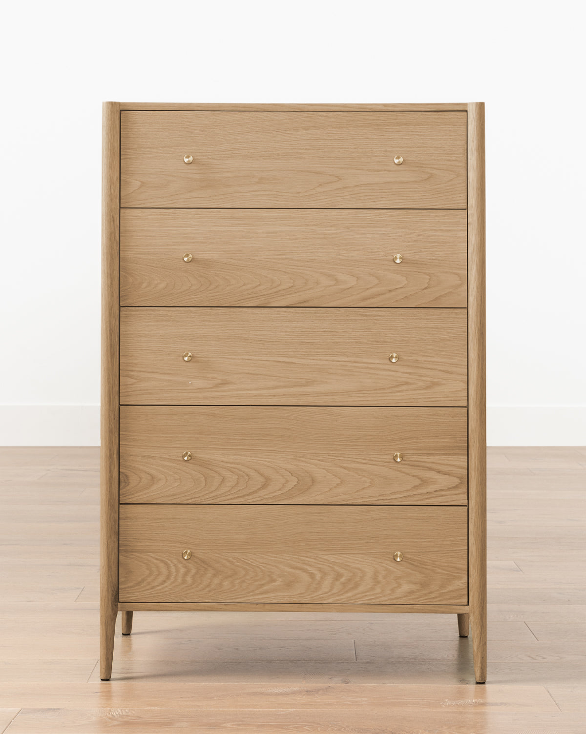 The Dana Tallboy Dresser showcases a clean, modern look in heritage white oak with five drawers and brass hardware, making it stand out beautifully on wood floors against a plain white wall, by McGee & Co.