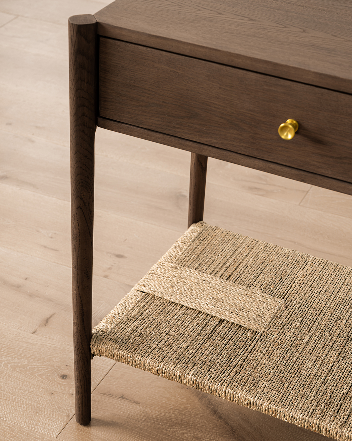 A close-up of the Dana 2-Drawer Console, showcasing its oak finish, round brass knobs, and woven lower shelf—ideal for entryway decor—set on a light wood floor, by McGee & Co.