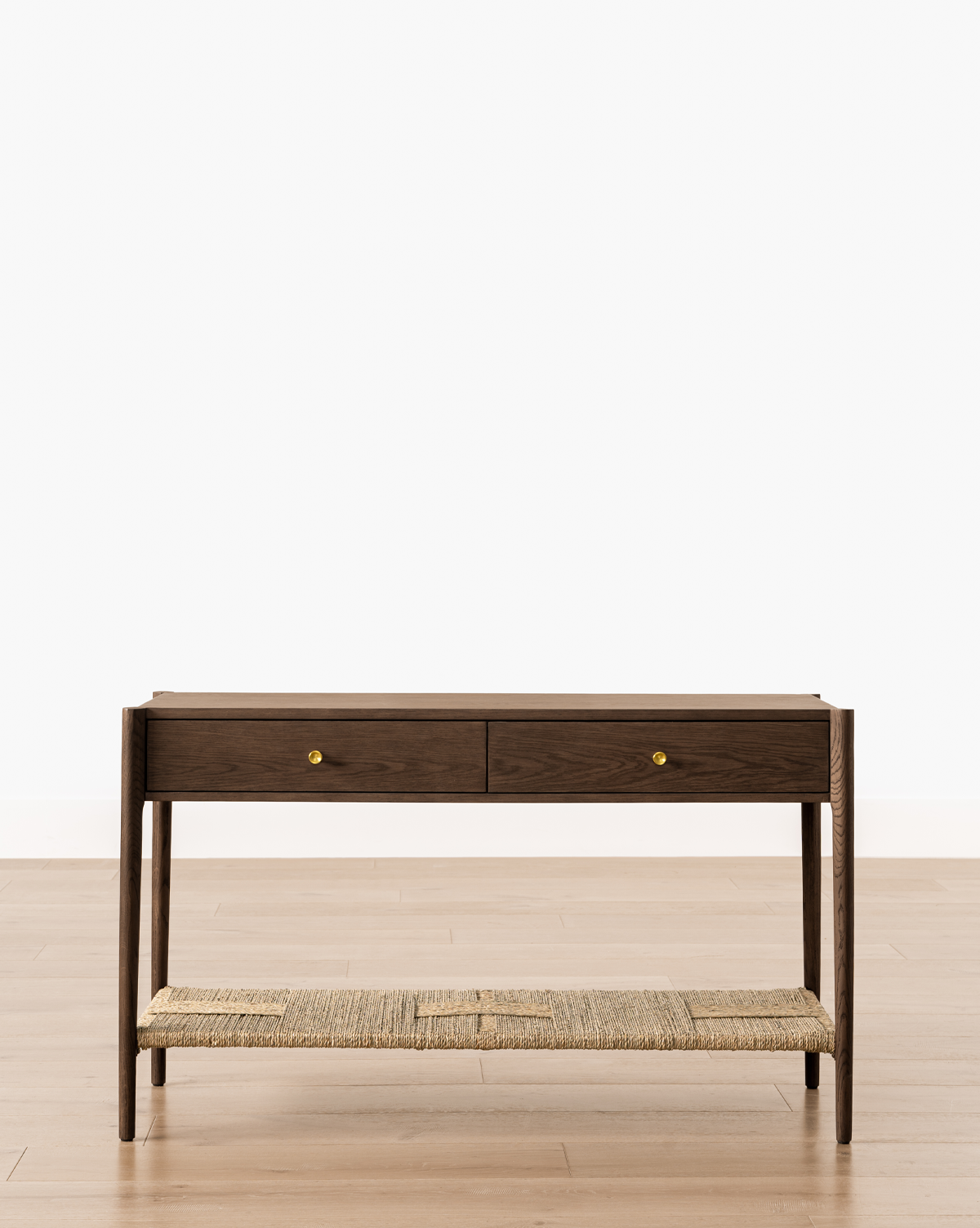 The Dana 2-Drawer Console, featuring gold knobs and a woven natural fiber lower shelf, stands on a light wood floor—perfect for entryway decor against a plain white wall, by McGee & Co.