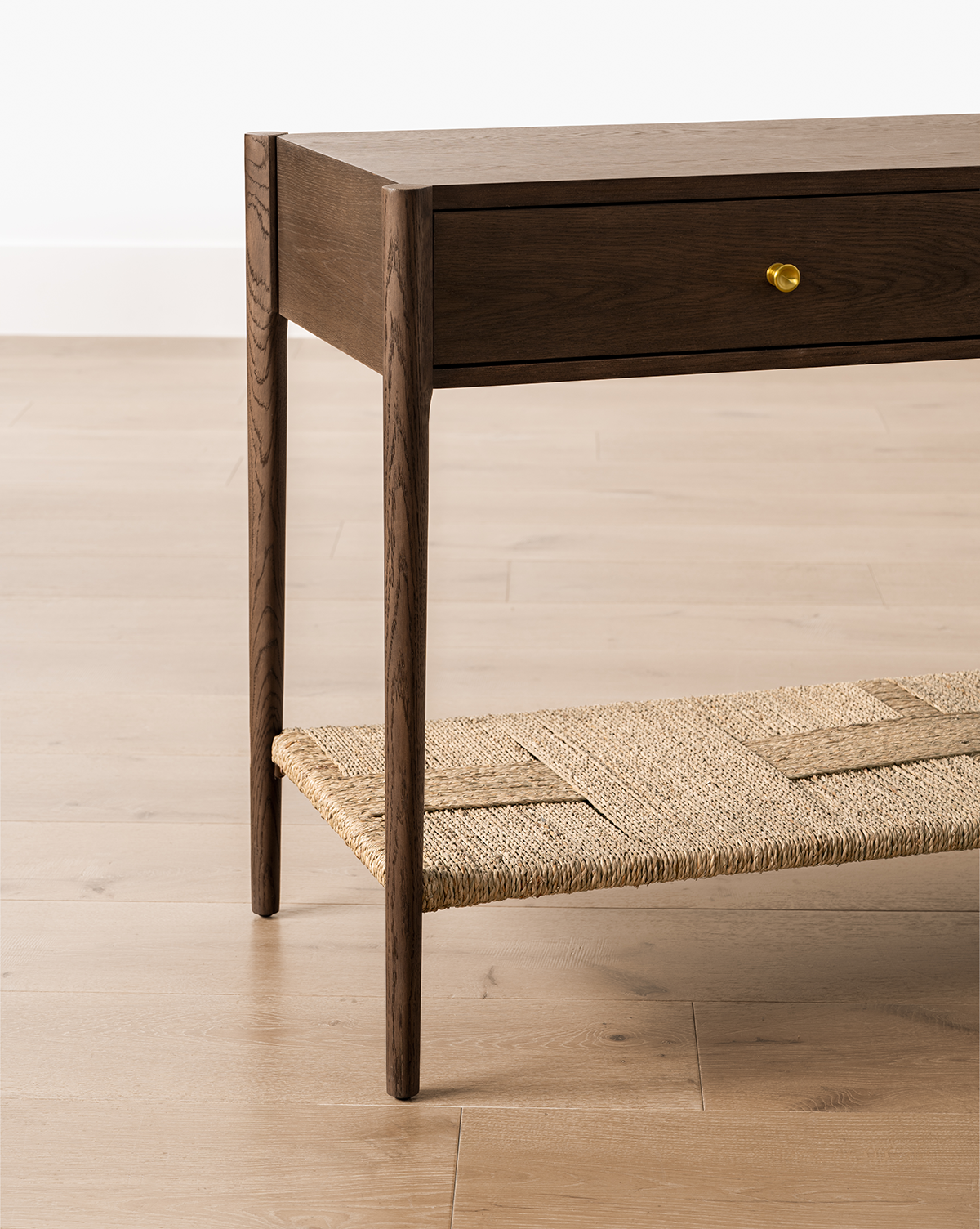 The Dana Walnut Oak Console, featuring slender legs, brass hardware on a single drawer, and a woven lower shelf, stands on a light wood floor against a white wall, by McGee & Co.