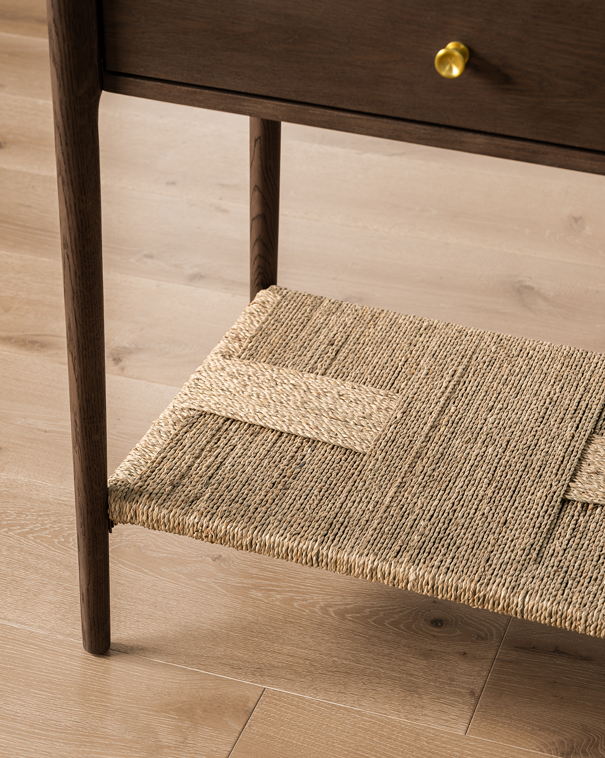 Close-up of the Dana Walnut Oak Console leg and woven shelf on a light wood floor. The console features a dark walnut oak finish and a drawer with elegant brass hardware, by McGee & Co.