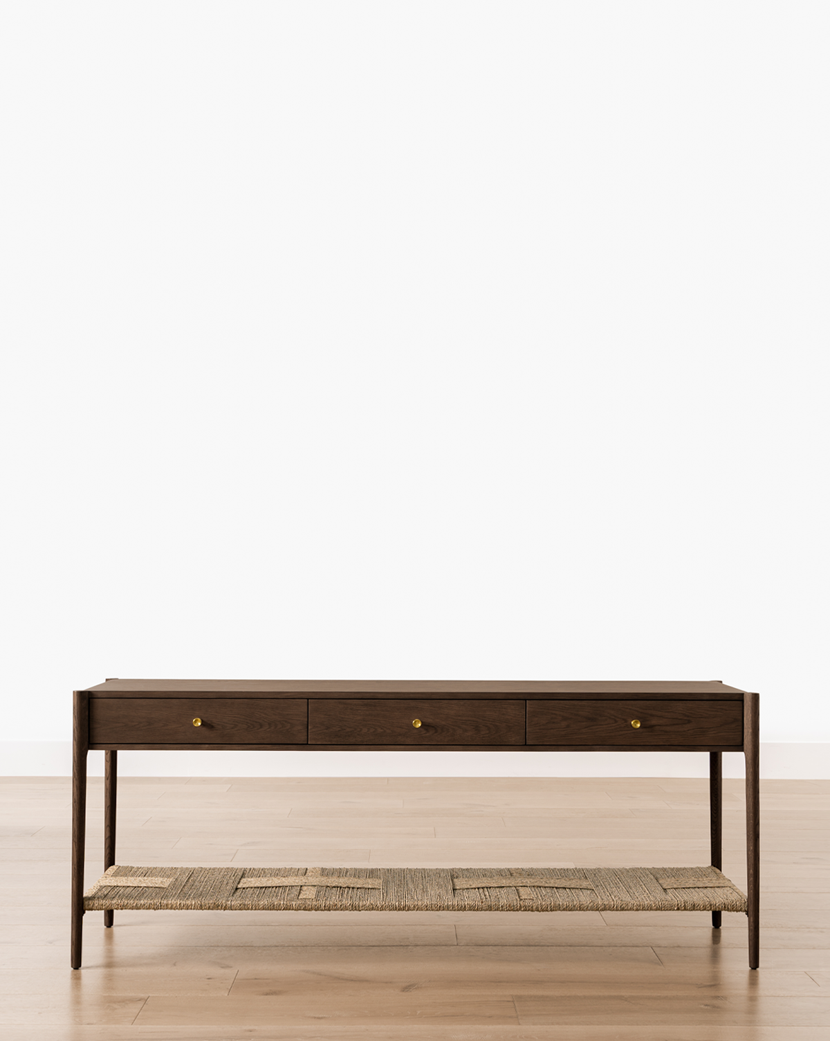 The Dana Walnut Oak Console, featuring three drawers, brass hardware, and a woven shelf, stands on a light wood floor against a plain white background, by McGee & Co.