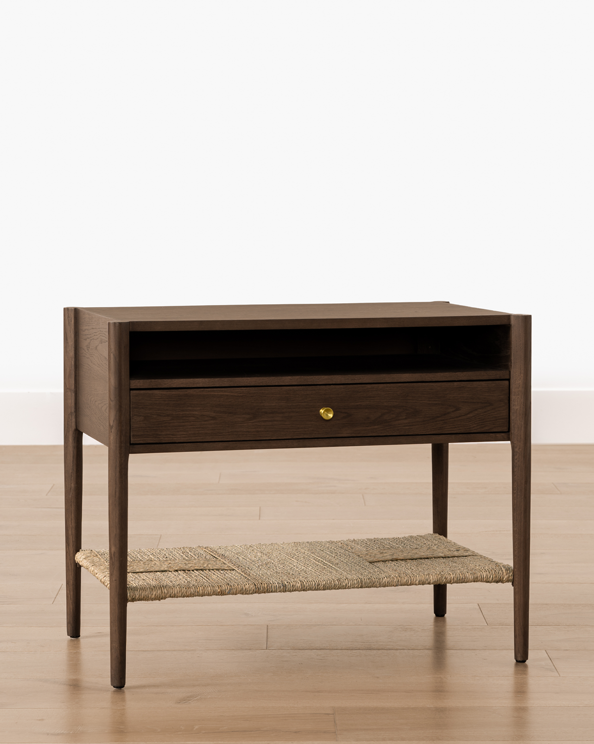The Dana Walnut Oak Nightstand features slender legs, a wide drawer with a brass knob, an open shelf above, and a woven rattan bottom shelf. It is shown on a light wood floor against a white background, by McGee & Co.
